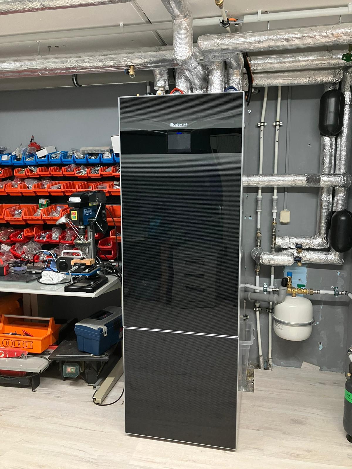 Schwarzer, großer Kühlschrank in Werkstatt mit Werkzeug im Hintergrund, umgeben von Rohren.