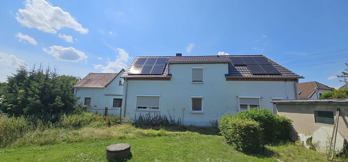 Ein weißes Haus mit Solarpanelen auf dem Dach an einem sonnigen Tag, umgeben von Gras und Büschen.