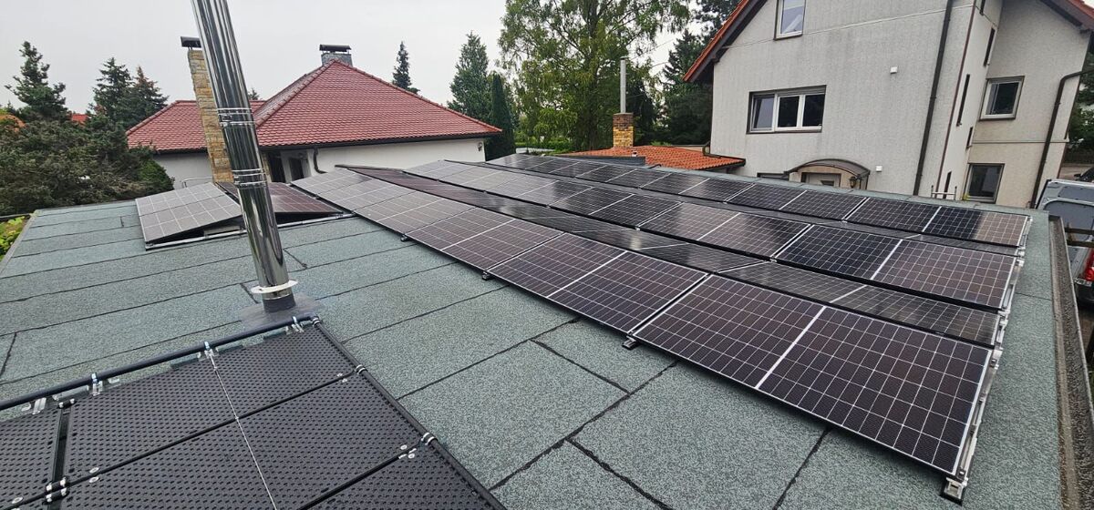 Solarmodule auf grünem Dach eines Hauses mit rotem Dach im Hintergrund, bewölkter Himmel.