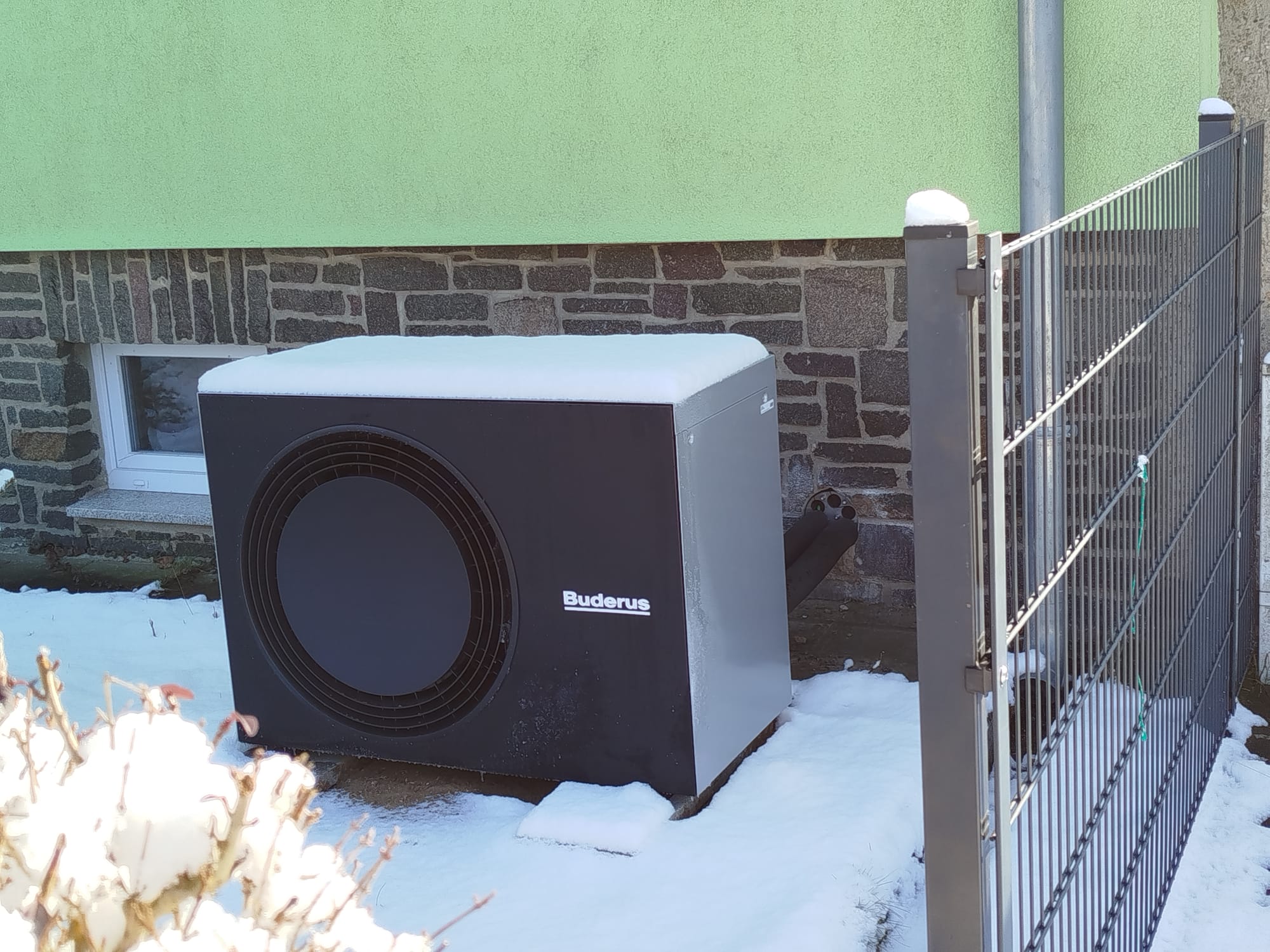 Wärmepumpe mit Schnee bedeckt, grüne und ziegelgraue Hintergrundwand, Zaun rechts sichtbar.