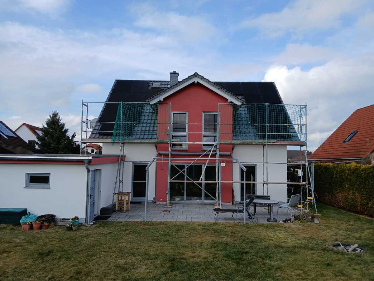 Einfamilienhaus mit rotem Giebel, Gerüst und grauen Dachschindeln unter bewölktem Himmel.