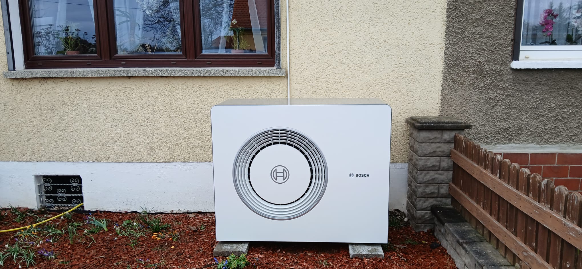 Klimaanlage an einer Außenwand, cremefarben, mit Bosch-Logo; steht auf zwei Betonblöcken vor Gebüsch.