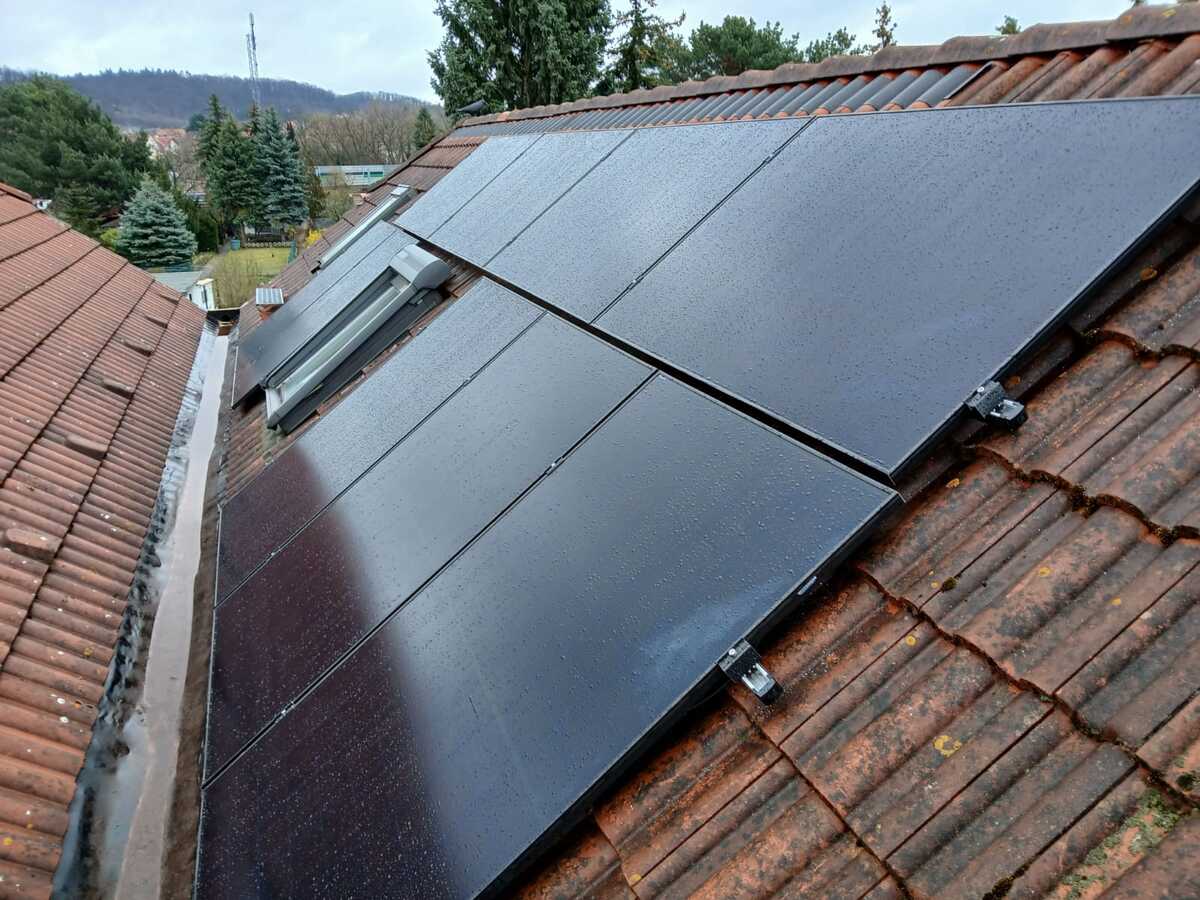 Solarmodule auf einem regennassen Ziegeldach an einem bewaldeten Hang, keine Menschen sichtbar.