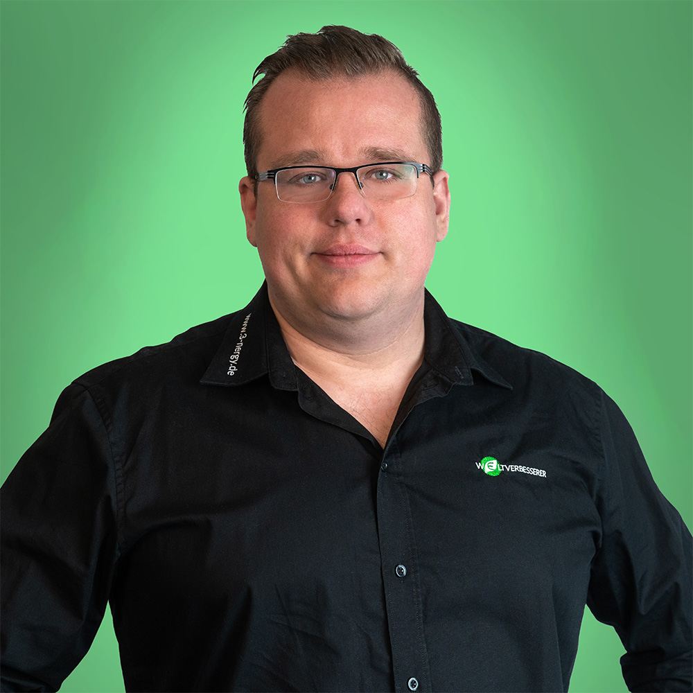 Ein Mann mit Brille lächelt, trägt ein schwarzes Hemd vor grünem Hintergrund.