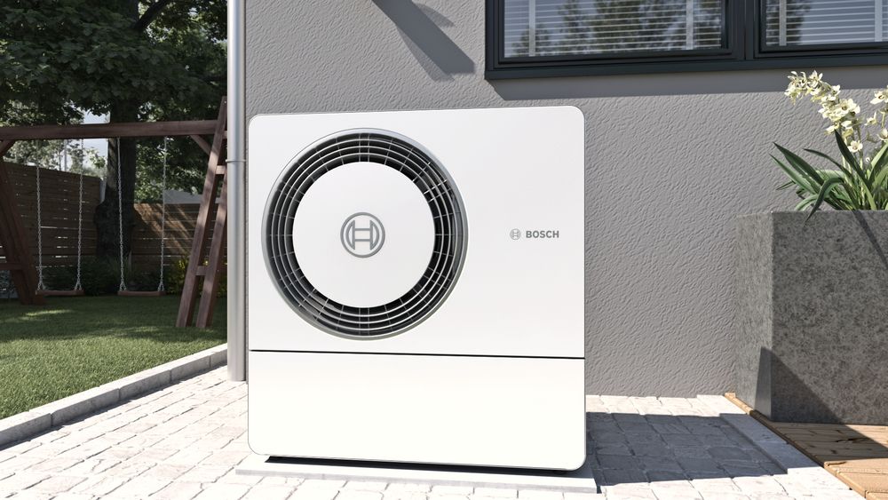 Weiße Bosch-Wärmepumpe im Freien, neben einem Gebäude, umgeben von Pflanzen und Kopfsteinpflaster.