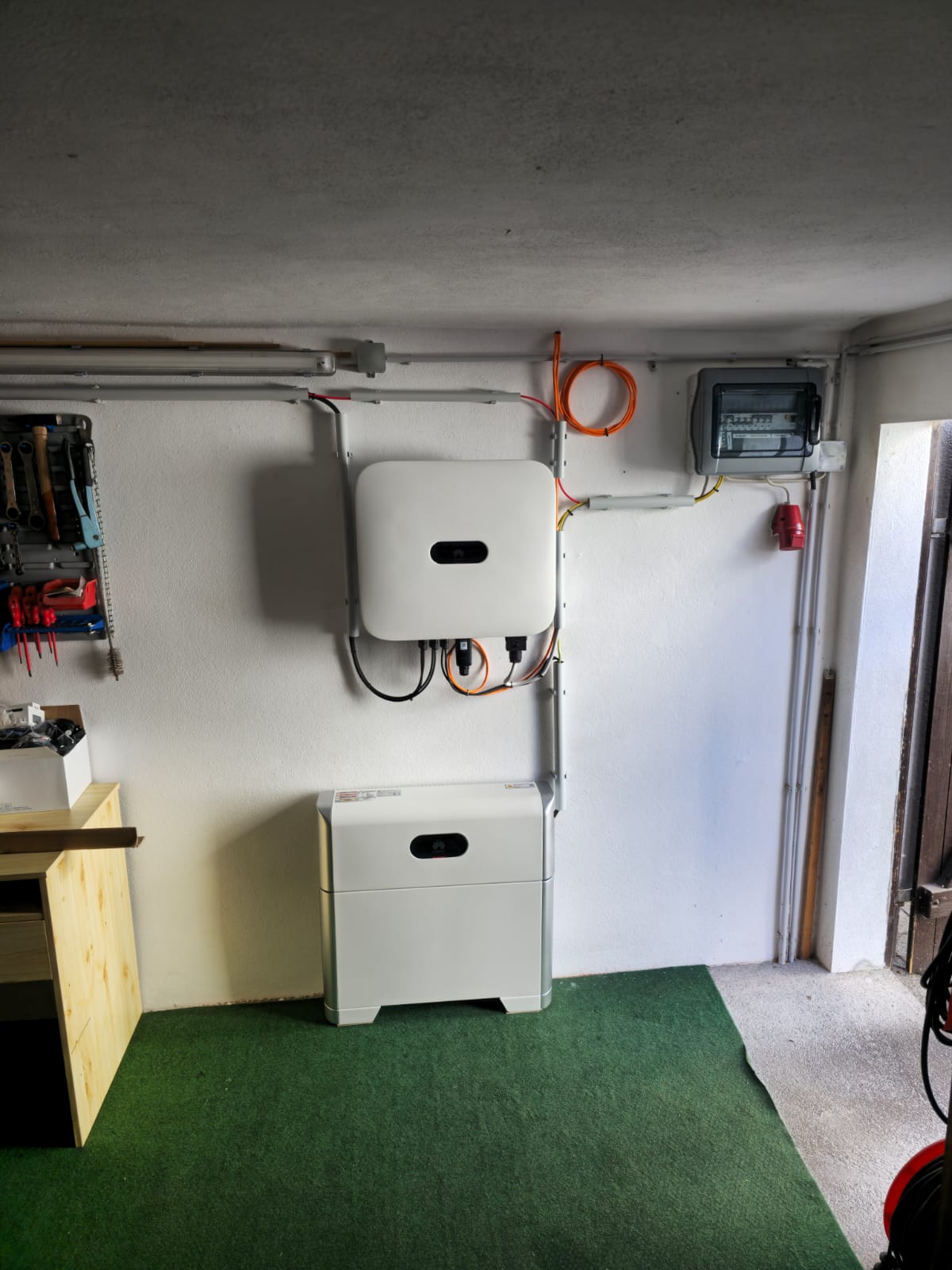Installierter Batteriespeicher mit Verkabelung an weißer Wand, grüne Bodenmatte.