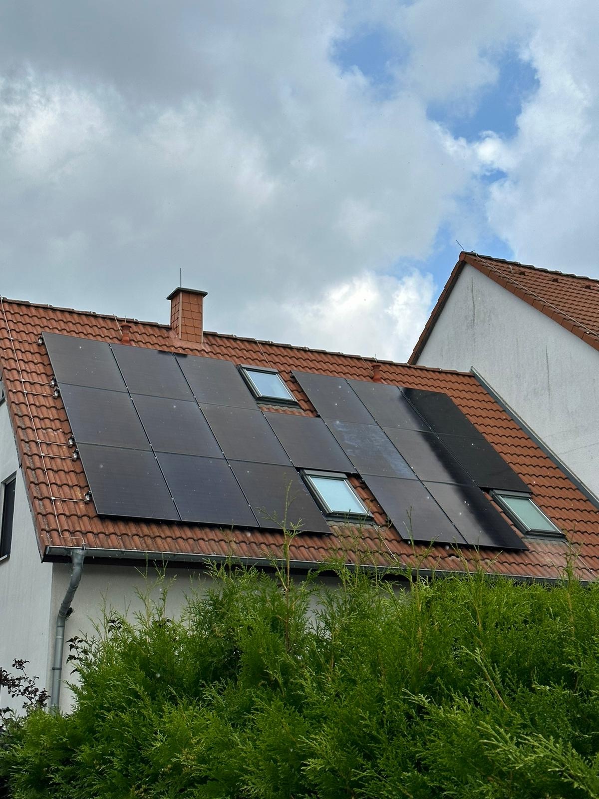 Ziegeldach mit Solarmodulen auf einem weißen Haus, umgeben von grünen Büschen und bewölktem Himmel.