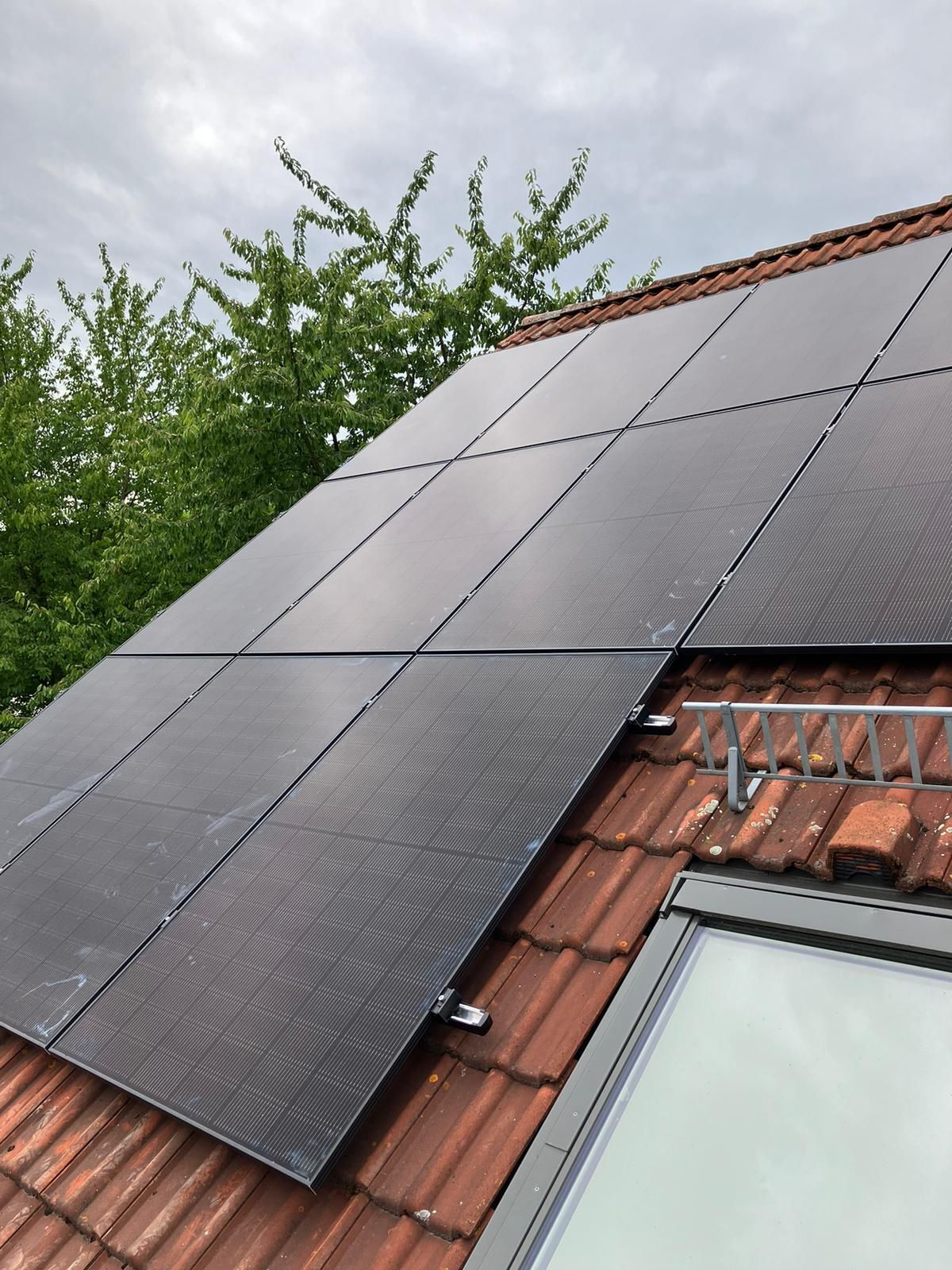 Solarmodule auf einem Ziegeldach, bewölkter Himmel, grüne Baumkronen im Hintergrund.