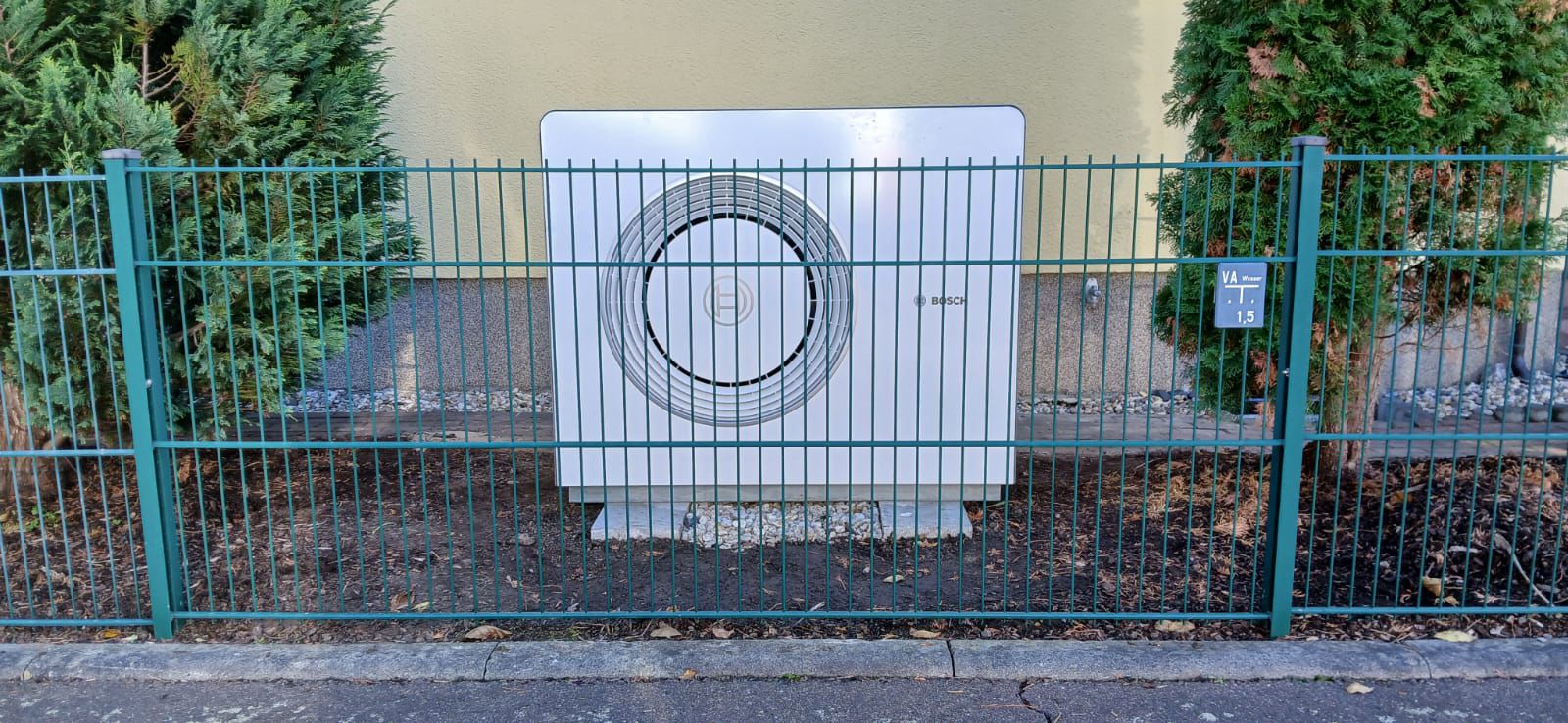 Eine Bosch-Außenklimaanlage hinter grünem Zaun zwischen zwei Bäumen, auf grauem Kies stehend.