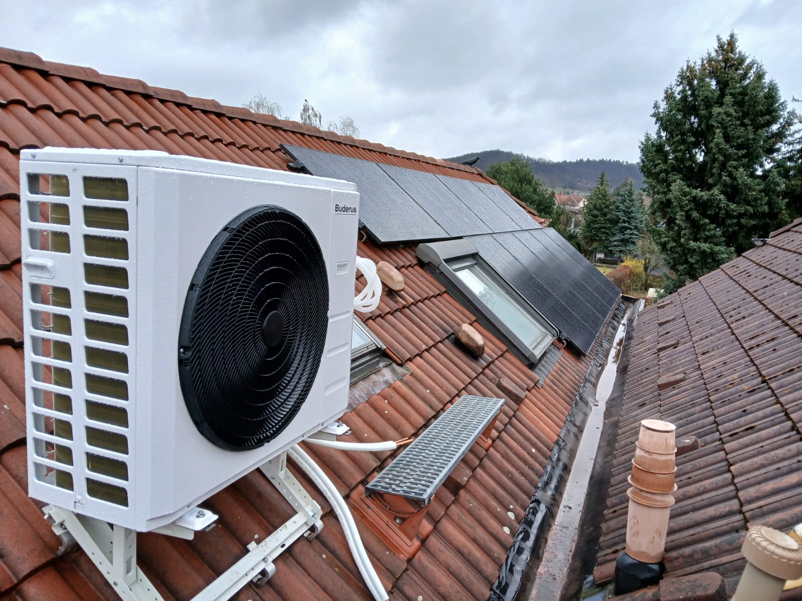 Klimaanlage und Solarpaneele auf einem roten Ziegeldach, bewölkter Himmel im Hintergrund.