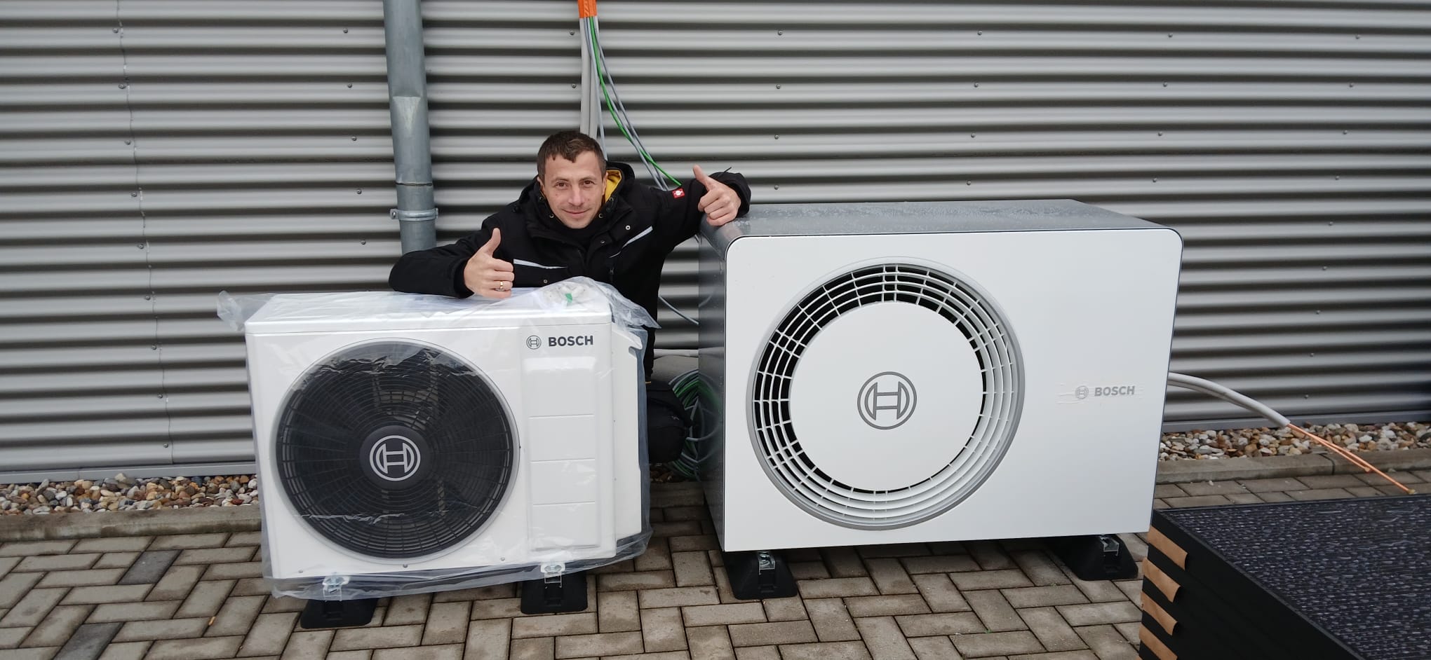 Eine Person lächelt, zeigt Daumen hoch; sitzt zwischen zwei Bosch-Klimaanlagen vor grauer Wand.
