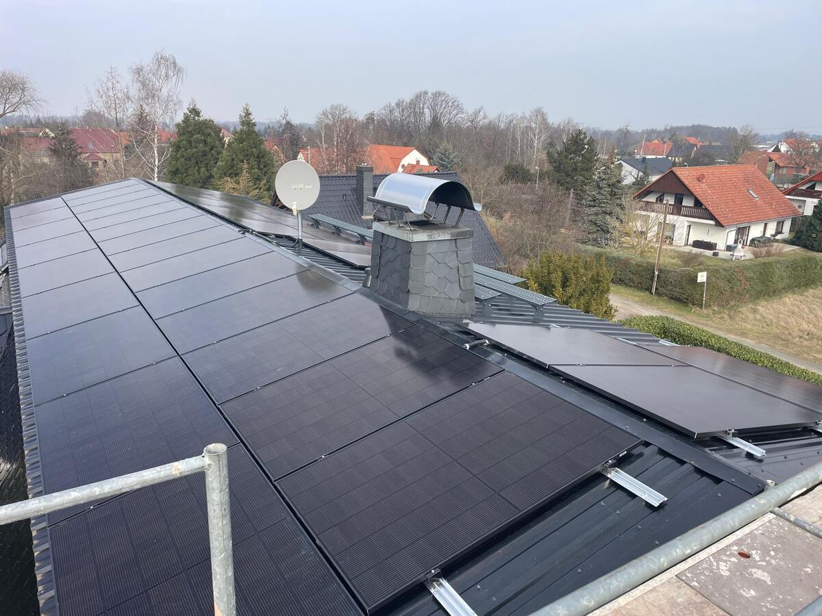 Dach mit schwarzen Solarpanels und Schornstein; Häuser und Bäume im Hintergrund.