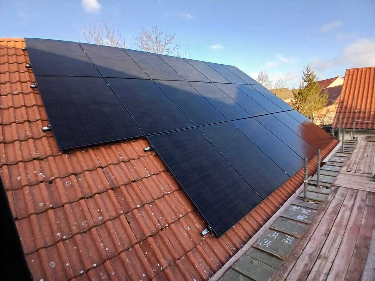 Auf einem roten Ziegeldach sind schwarze Solarmodule montiert; blauer Himmel im Hintergrund.