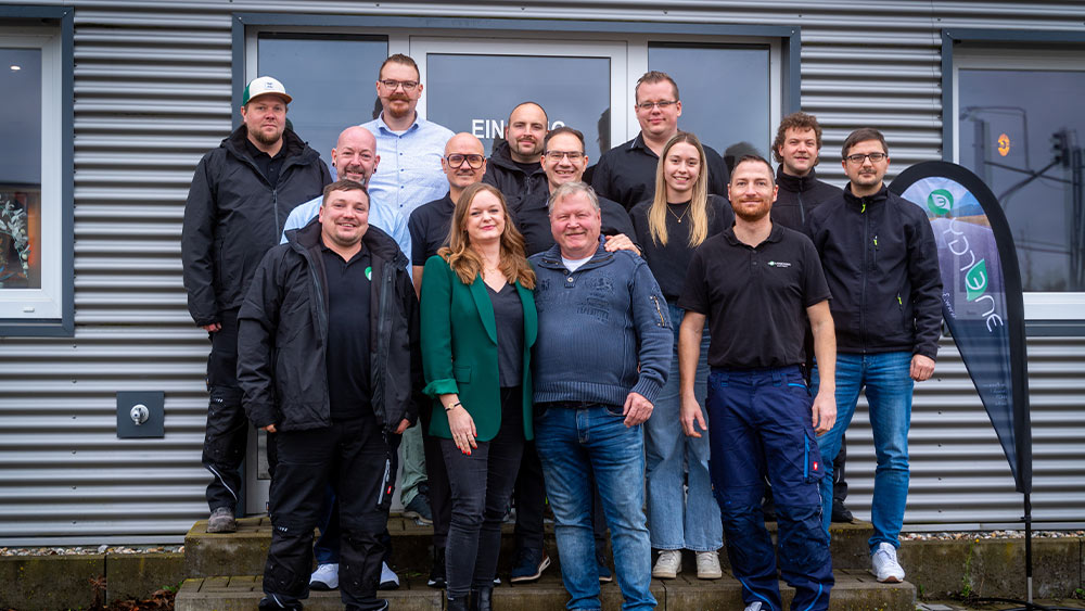 Eine Gruppe von 14 Personen posiert lächelnd vor einem Metallgebäude in Arbeitskleidung.