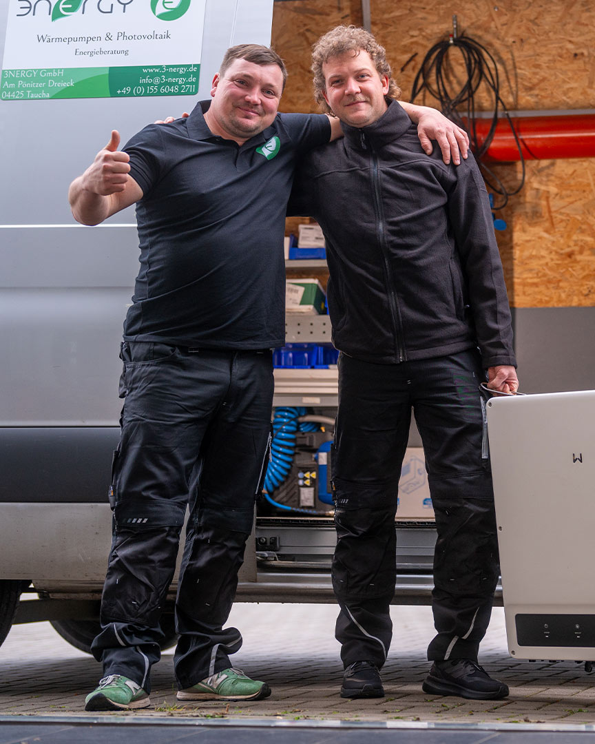 Zwei Männer in Arbeitskleidung posieren lächelnd vor einem Transporter in einer Werkstatt.