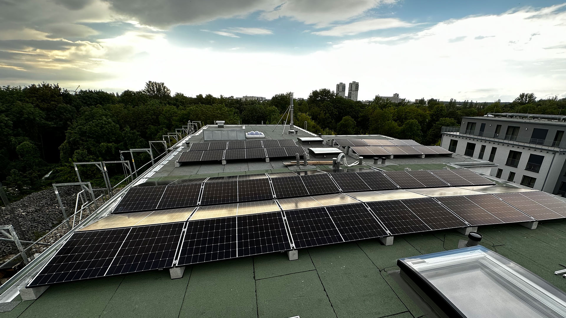 Dach mit Solarmodulen bei bewölktem Himmel; grüne Umgebung und moderne Gebäude im Hintergrund.
