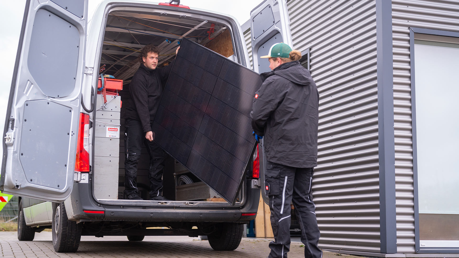 Zwei Männer verladen ein großes Solarpanel in einen Lieferwagen; konzentrierte Gesichtsausdrücke.