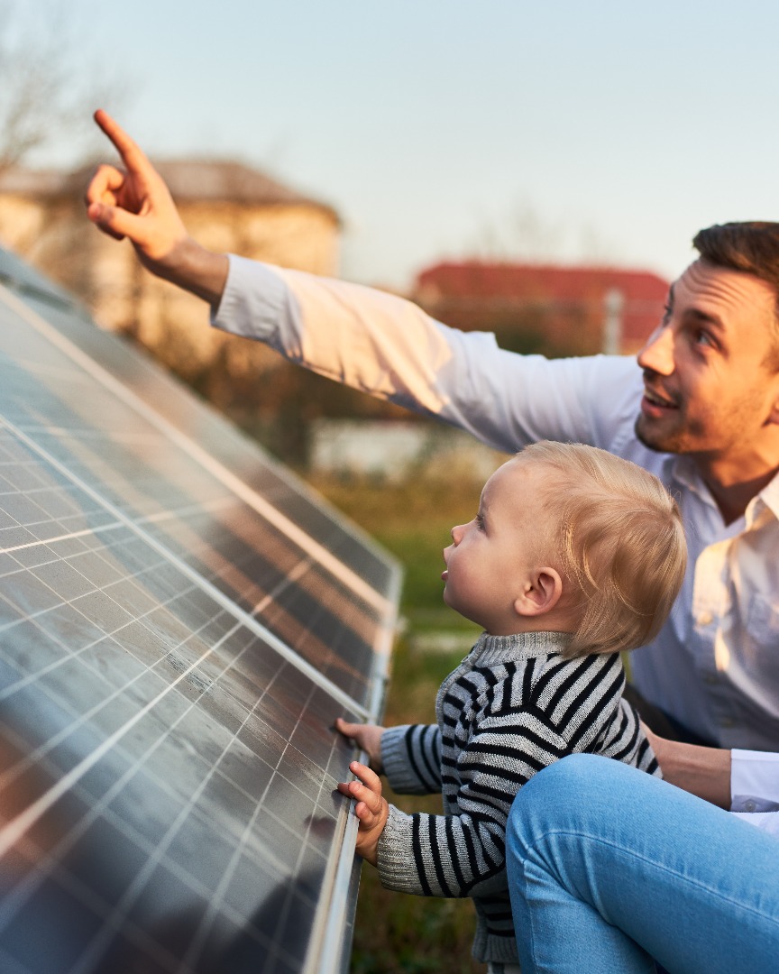 Man shows his family the solar panels on the plot near the house during a warm day. Young woman with a kid and a man in the sun rays look at the solar panels.