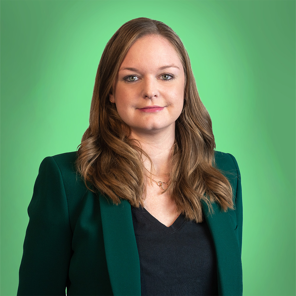 Eine Frau mit braunem Haar lächelt leicht. Sie trägt einen grünen Blazer vor grünem Hintergrund.