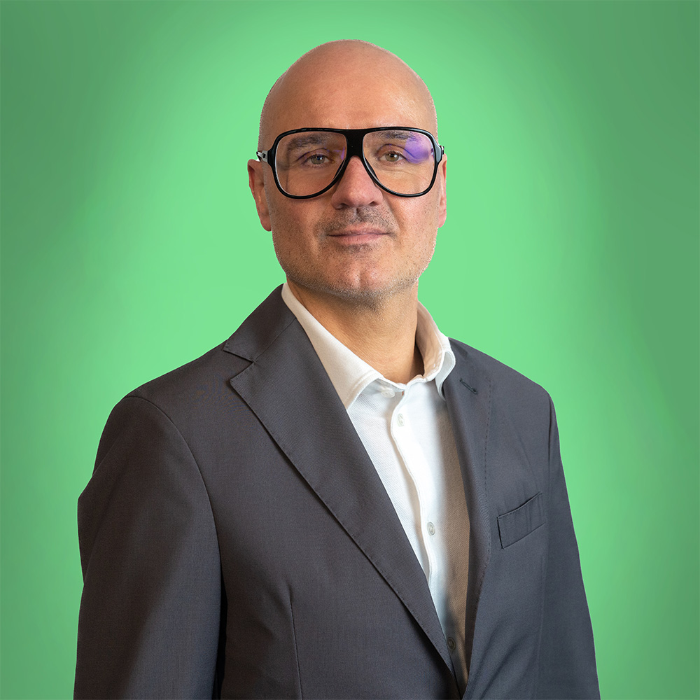 Mann mit Glatze in Anzug, großer Brille, ernster Blick; Grüner Hintergrund.