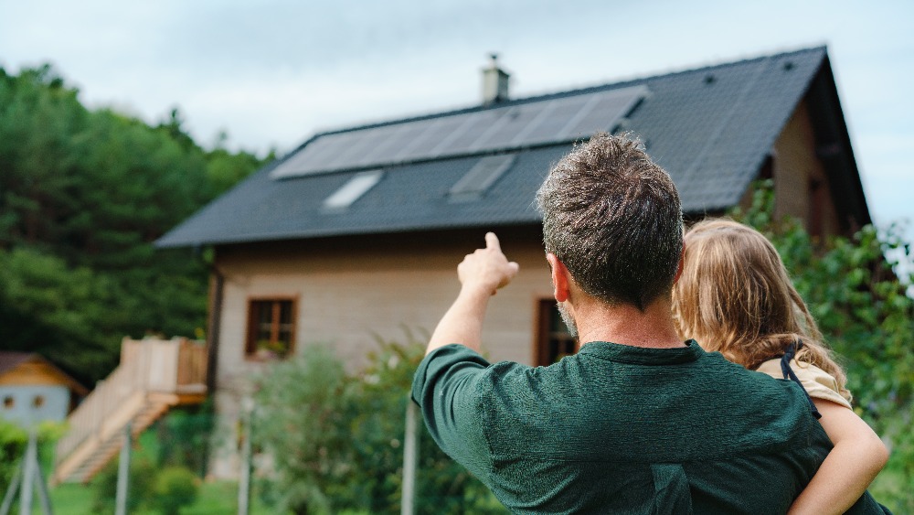 Rear view of dad holding her little girl in arms and showing at their house with installed solar panels. Alternative energy, saving resources and sustainable lifestyle concept.