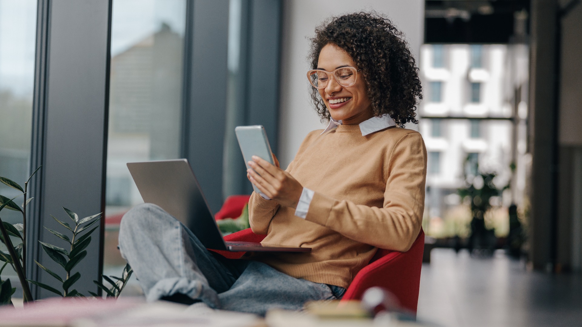 Smiling business woman is using phone sitting in office during break time with laptop